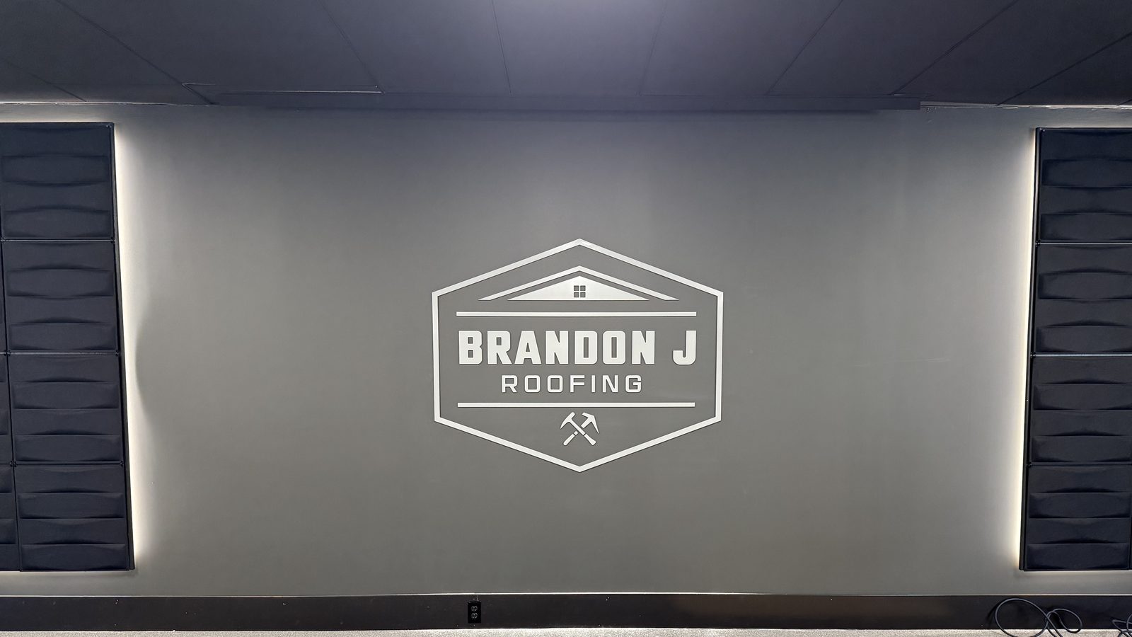 Gray Brandon J Roofing logo badge cut from PVC, flush mounted on a dark gray office wall between two backlit acoustic panels